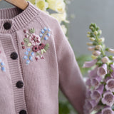 Pink knitted cardigan with floral embroidery and black buttons, hanging against a natural background with flowers.