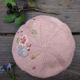 Pink knitted beret with floral embroidery on a wooden surface with blackberries and leaves.