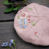 Pink knitted beret with floral embroidery on a wooden surface with flowers.