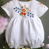 White baby romper with floral embroidery on a wooden hanger surrounded by flowers.