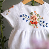 White baby romper with floral embroidery on a wooden hanger against a natural background.