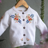 White knitted cardigan with floral embroidery on a hanger against a natural background.