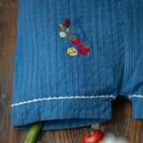 Blue fabric with floral embroidery on a wooden surface with vegetables and flowers.