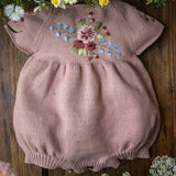 Pink baby romper with floral embroidery on a wooden surface surrounded by flowers