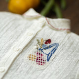 White garment with embroidered design of a person holding a bag of groceries, surrounded by fruits on a wooden surface.