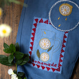 Embroidered blue fabric with a lemon and cup design on a wooden surface with flowers.