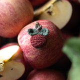 A close-up of red apples, including whole apples and cut apple halves, with a small crocheted raspberry ornament attached to one apple.