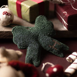 A dark green beaded bow ornament lies on a dark red surface, surrounded by festive props including wrapped gifts, candy canes, and a plush toy in a moody holiday setting.