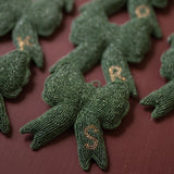 A close-up of several dark green beaded bow ornaments with gold embroidered letters, arranged on a dark red background.