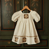 White dress with brown floral patterns hanging on a wooden hanger against a dark green wall with decorative patterns.