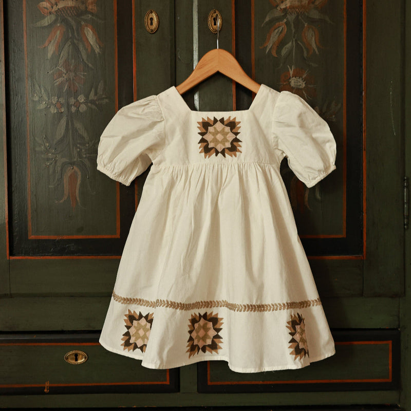 White dress with brown floral patterns hanging on a wooden hanger against a dark green wall with decorative patterns.