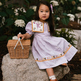 Girl wearing the Birdie Bliss Dress - Lilac, surrounded by flowers and a basket.