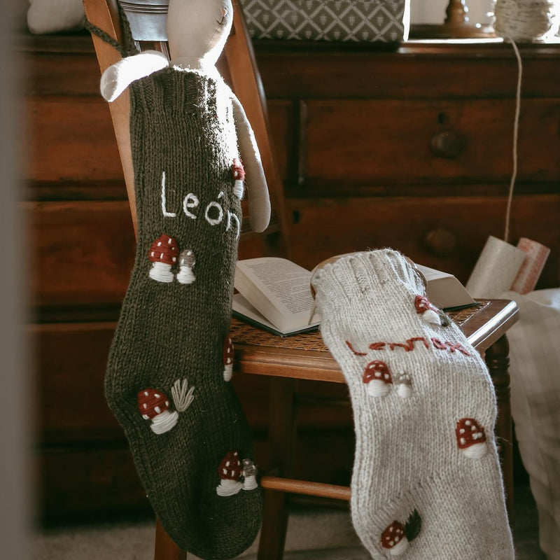 Two moss-colored personalized wool stockings with hand-embroidered mushroom patterns, displayed on different parts of a chair.