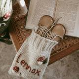 Close-up of a rye-colored personalized wool stocking with a hand-embroidered mushroom pattern, partially visible and holding two small shoes at the top, surrounded by Christmas decorations.