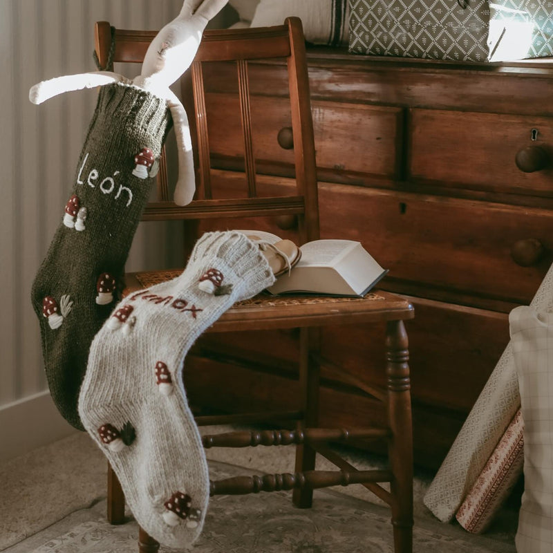  Two rye-colored personalized wool stockings with hand-embroidered mushroom patterns, displayed on different parts of a chair.