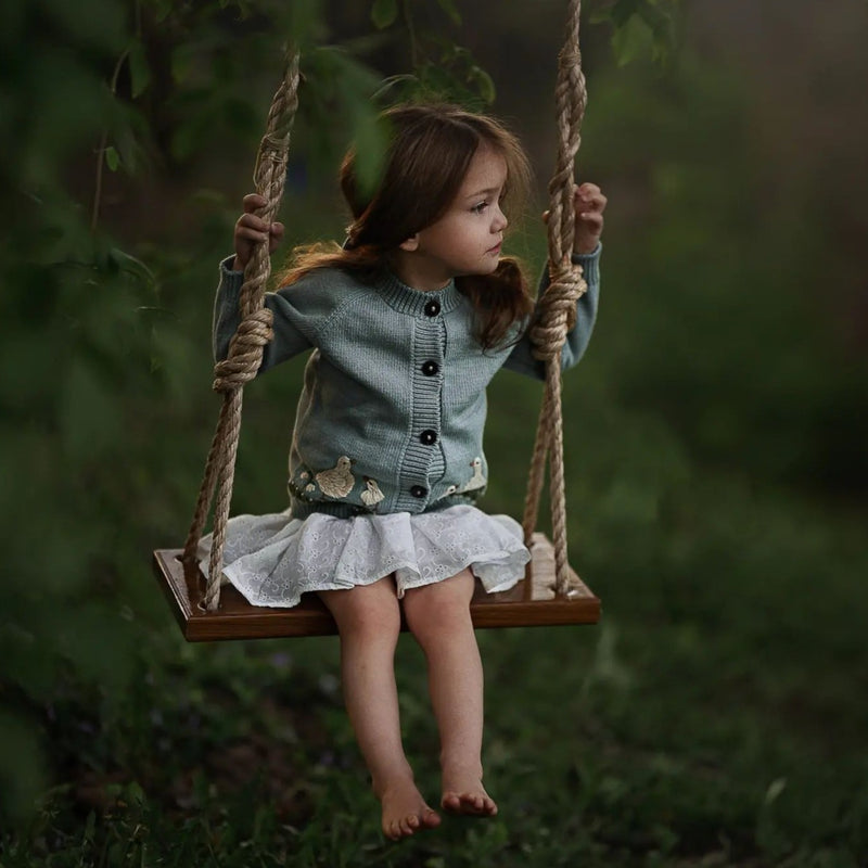 A girl on a swing wearing the Spring Geese Cardigan in Duck Blue, surrounded by natural greenery, creating a peaceful, springtime scene.

