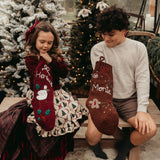Two children with personalized Christmas stockings in a festive setting with decorated trees.