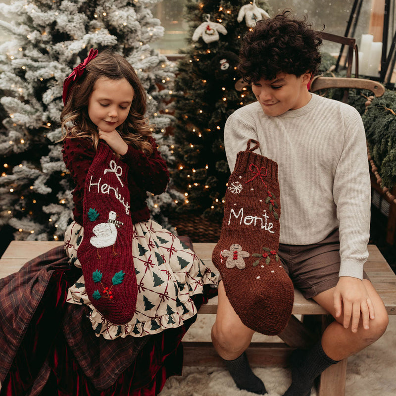 Two children with personalized Christmas stockings in a festive setting with decorated trees.