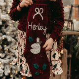 Child wearing a burgundy sweater with a name on it, standing in front of a decorated Christmas tree.