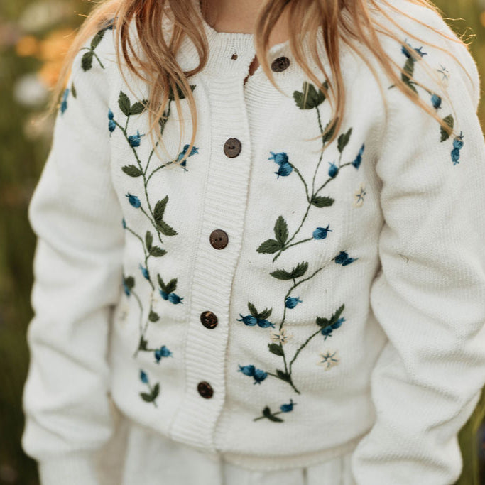 White cardigan with blueberries embroidery worn by a person in a field of flowers