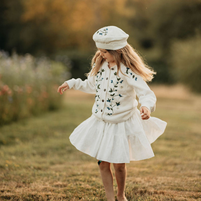 Young girl in a white dress and hat standing in a field with trees in the background