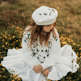 Young girl in a white dress with floral patterns and a matching beret sitting in a field of yellow flowers.