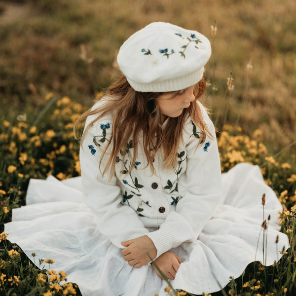 Young girl in a white dress with floral patterns and a matching beret sitting in a field of yellow flowers.