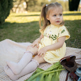 Young girl in a yellow dress sitting on a blanket with flowers and a bag in a park.