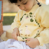 Child wearing a yellow sweater with a nature-themed design, sitting outdoors.