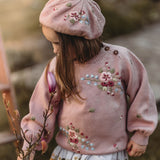 Little girl wearing a pink embroidered sweater and matching beret outdoors
