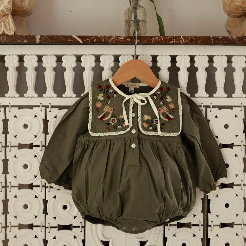 Green baby romper with floral embroidery on a hanger against a decorative wall.