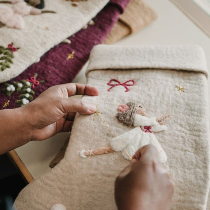 Artisan hand-knitting a cream white holiday stocking, focusing on the creation of the embroidered Odette and holiday motifs, showcasing skilled craftsmanship.