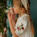 Young girl wearing a white dress with strawberry embroidery and a lace headband, standing next to greenery.