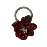A handmade beaded flower napkin ring with a large red and green beaded flower attached to a sage green braided elastic loop, shown on a plain white background.