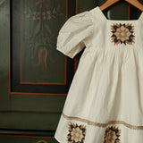 White dress with floral embroidery on a wooden hanger against a green wall with decorative patterns.