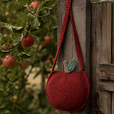 A crocheted red apple-shaped shoulder bag with a green leaf accent hangs on a rustic wooden fence post in an apple orchard.