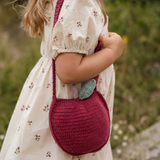 A child wearing a cream floral dress carries a crocheted apple-shaped shoulder bag in deep pink with a green leaf accent, set against a blurred outdoor background.