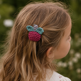 A young girl with long light brown hair is shown in profile outdoors, wearing a crocheted hair accessory shaped like a raspberry with teal leaves and a small butterfly on a string.