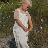 Child in a field of wildflowers wearing a beige top and white overalls with floral patterns.
