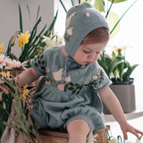 Baby in a light blue outfit with animal patterns sitting on a chair surrounded by flowers.