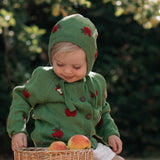 A toddler wearing a green knit hooded cardigan with red apple prints sits on a wooden stool outdoors, holding a wicker basket with apples. The background is a soft-focus garden with green foliage.