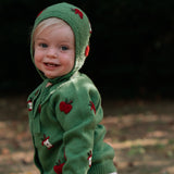 A toddler standing outdoors wearing a green knit hooded cardigan and matching bonnet with red apple motifs, paired with white textured diaper cover.