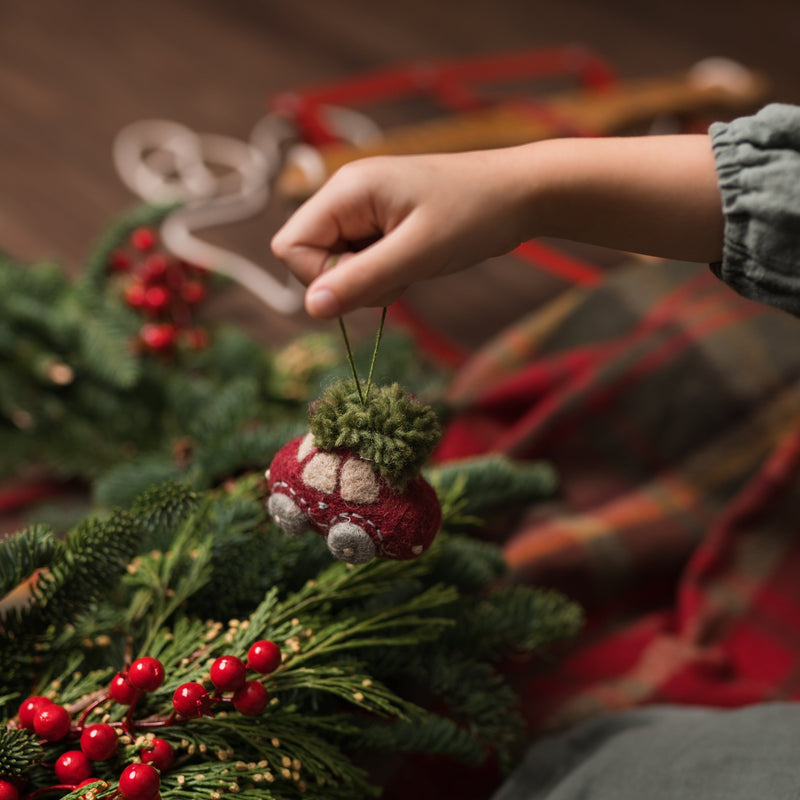 Car Ornament with a vintage car and Christmas tree, held in a hand with festive greenery or a decorative tree branch visible beneath it