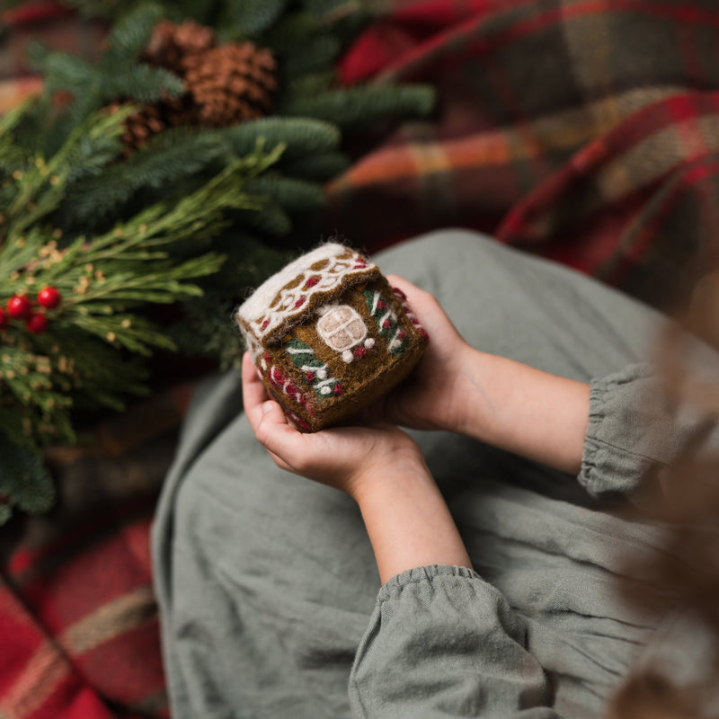 Handcrafted Gingerbread House Ornament in brown, held in hands with blurred festive greenery in the background











