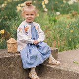 Young girl in a floral cardigan and blue dress sitting on steps with flowers in the background