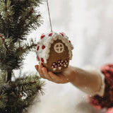 Hand holding a gingerbread house ornament against a blurred Christmas tree background