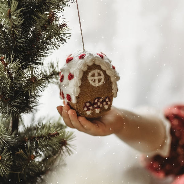 Hand holding a gingerbread house ornament against a blurred Christmas tree background