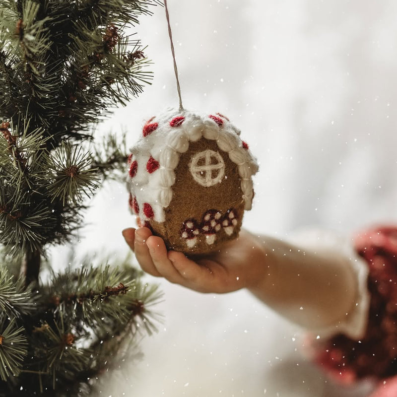 Hand holding a gingerbread house ornament against a blurred Christmas tree background