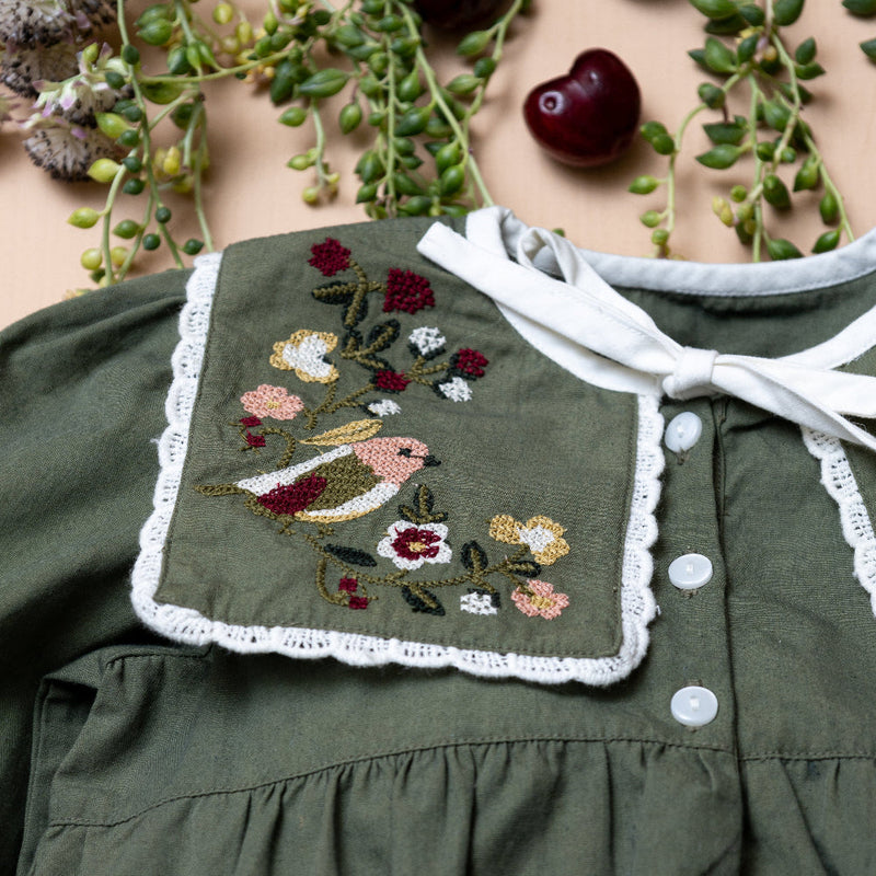 close up of green sewn romper with embroidered collar on a beige background with plants and fruit