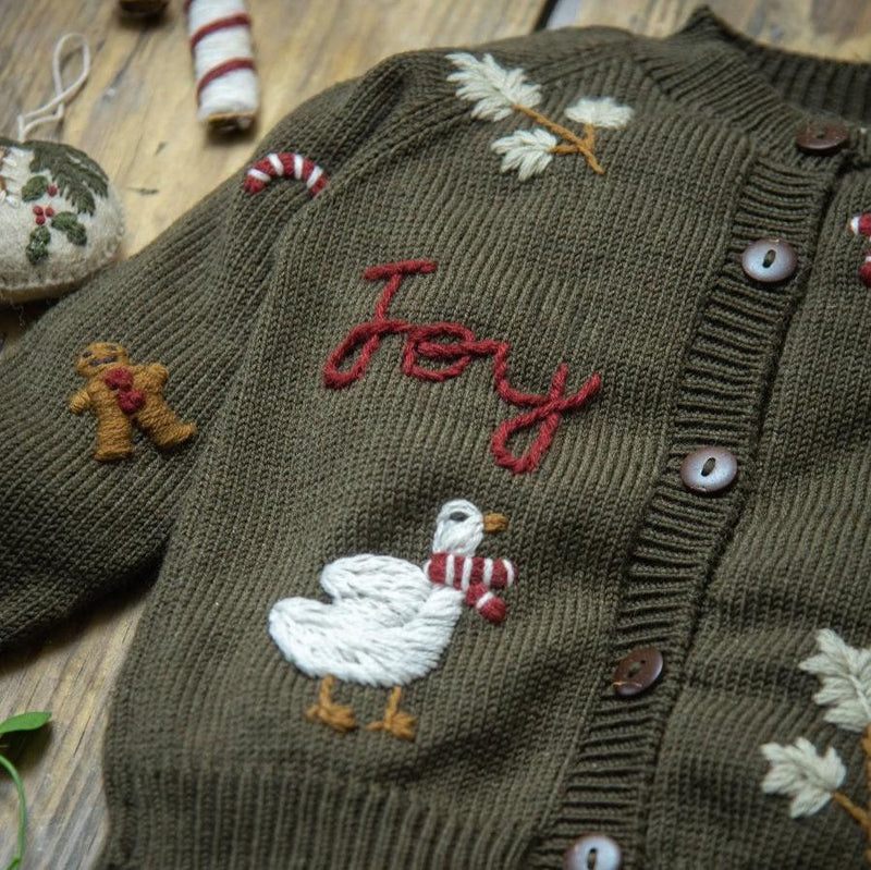 Close-up of the pine green Joy Cardigan, highlighting the intricate hand-embroidered holiday motifs including candy canes, gingerbread men, and festive geese.
